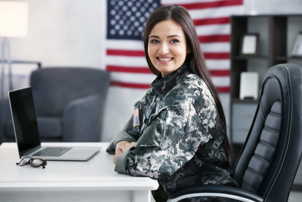 Female solder smiling at the camera, sitting at a desk with a laptop