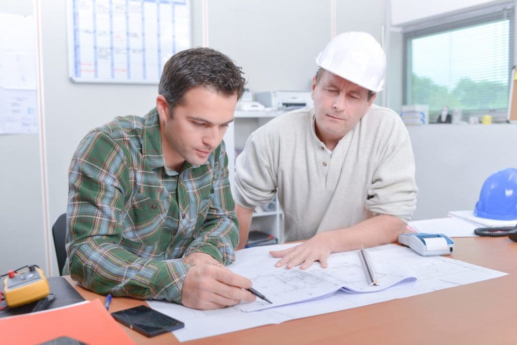 Two gentlemen, one on right wearing a hard hat, reviewing blueprints sitting at a desk