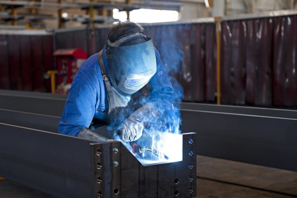 Welder works on metal fabrication, fumes billow up into his face which is protected by a mask